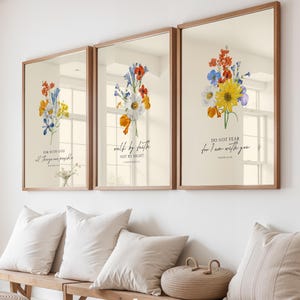 May include: Three framed floral art prints with inspirational quotes. Each print features a bouquet of colorful flowers in shades of yellow, blue, and orange. The frames are a light brown color, and the prints are displayed above a wooden bench with pillows.