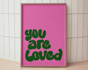 May include: A pink framed poster with the text "you are loved" in green bubble letters. The poster is on a white tiled wall.