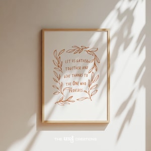 May include: A framed print with the text "LET US GATHER TOGETHER AND GIVE THANKS TO THE ONE WHO PROVIDES." The text is in a brown script font, surrounded by a wreath of leaves. The frame is a light wood color.