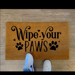 Könnte beinhalten: Eine rechteckige Fußmatte aus natürlicher brauner Kokosfaser. Die Matte zeigt den Schriftzug "Wipe your PAWS" in schwarzer Schrift und Pfotenabdruck-Grafiken. Weiße Turnschuhe sind am unteren Bildrand zu sehen.