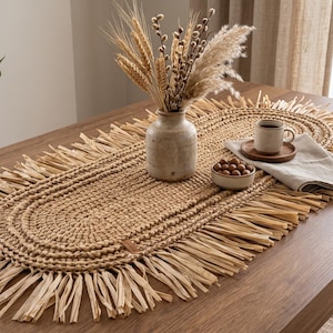 May include: An oval-shaped, woven tan table runner featuring a thick, braided center and long, fringed edges. The runner is placed on a wooden table and holds a ceramic vase with dried wheat, a small bowl of nuts, and a coffee mug on a napkin.
