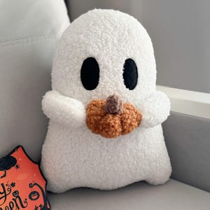 Könnte beinhalten: Ein weißes Plüschgespenst mit einer strukturierten, flauschigen Oberfläche, zwei schwarzen ovalen Augen und kleinen Armen, die einen kleinen, runden, orangefarbenen Kürbis mit braunem Stiel halten. Daneben steht ein Schild mit dem Text Happy Halloween.