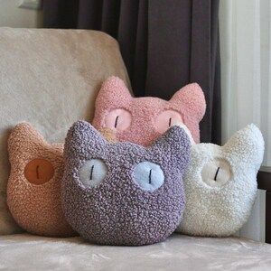 May include: A collection of four cat-shaped throw pillows made of textured, fuzzy sherpa fabric. The pillows are colored in shades of dusty pink, light brown, muted purple, and cream, each featuring two embroidered eyes with vertical black pupils.