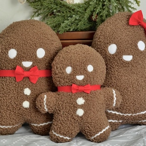 Puede incluir: Tres cojines de felpa con forma de hombre de jengibre de diferentes tamaños. Son marrones con detalles blancos, incluyendo ojos, sonrisas y detalles de glaseado. Cada uno tiene un lazo rojo. Los cojines están sobre una cama con una manta gris y blanca estampada.