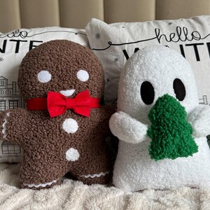 May include: A brown gingerbread person plush with white eyes and buttons, wearing a red bow tie, next to a white ghost plush holding a green textured tree. Both are made of a fuzzy, sherpa-like fabric, positioned in front of white pillows with the text hello WINTER.