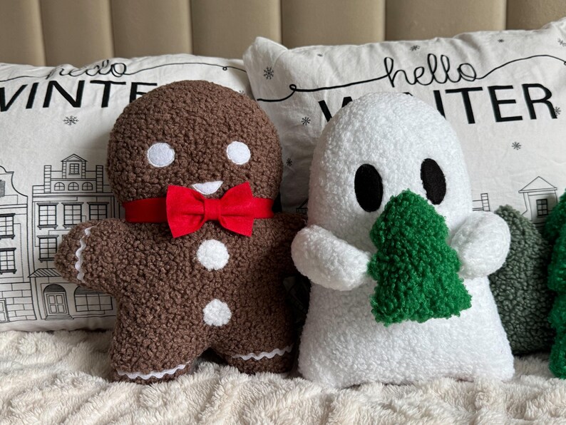 May include: A brown gingerbread person plush with white eyes and buttons, wearing a red bow tie, next to a white ghost plush holding a green textured tree. Both are made of a fuzzy, sherpa-like fabric, positioned in front of white pillows with the text hello WINTER.
