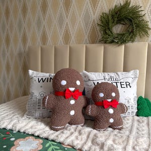 May include: Two brown textured gingerbread man plush toys with white circular eyes, white buttons, and red bow ties. The plush toys are positioned in front of two white pillows featuring the text hello WINTER and a black line drawing of houses.