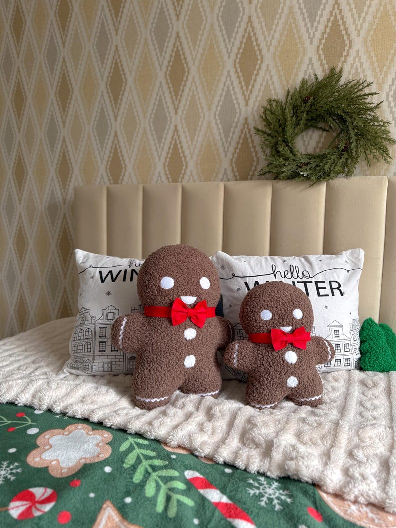 May include: Two brown textured gingerbread man plush toys with white circular eyes, white buttons, and red bow ties. The plush toys are positioned in front of two white pillows featuring the text hello WINTER and a black line drawing of houses.