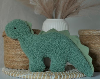May include: A sage green dinosaur plush toy made of textured sherpa fabric. The dinosaur features a small black eye and a smooth, light green fabric spine that runs from the back of the head down to the tip of the tail. The body has a rounded head, four short legs, and a long, pointed tail.