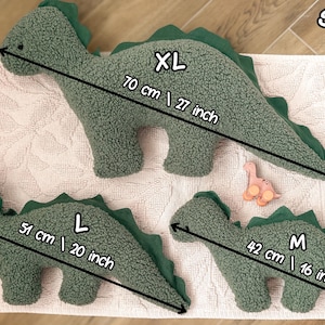 May include: Three sage green dinosaur-shaped plush pillows with textured fabric and dark green felt spines. The XL size measures 27 inches, the L size measures 20 inches, and the M size measures 16 inches in length. A small pink toy is also visible.