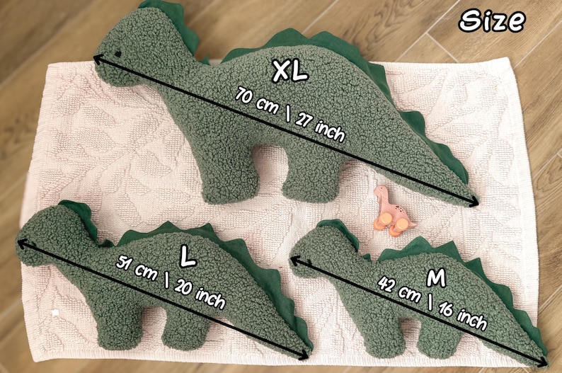 May include: Three sage green dinosaur-shaped plush pillows with textured fabric and dark green felt spines. The XL size measures 27 inches, the L size measures 20 inches, and the M size measures 16 inches in length. A small pink toy is also visible.
