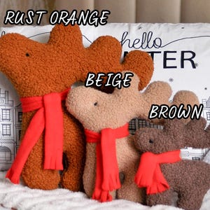 May include: Three plush reindeer toys in rust orange, beige, and brown, each featuring a textured fabric surface and a bright red scarf. The toys are labeled with the text RUST ORANGE, BEIGE, and BROWN, and are positioned in front of a pillow.