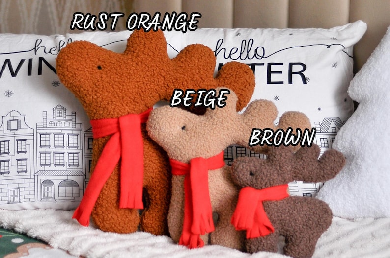 May include: Three plush reindeer toys in rust orange, beige, and brown, each featuring a textured fabric surface and a bright red scarf. The toys are labeled with the text RUST ORANGE, BEIGE, and BROWN, and are positioned in front of a pillow.