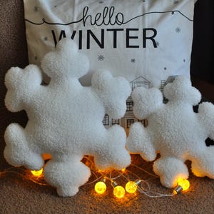 May include: Two white plush snowflake pillows with a white pillow with the text "hello winter" behind them. The pillows are surrounded by string lights.