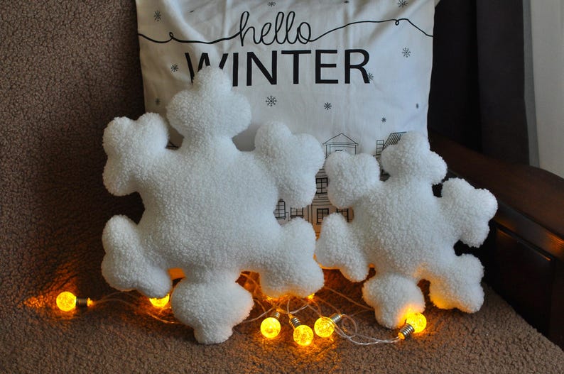 May include: Two white plush snowflake pillows with a white pillow with the text "hello winter" behind them. The pillows are surrounded by string lights.