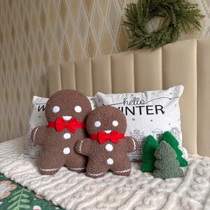 May include: Two brown textured gingerbread man plushies with white circular eyes and buttons, wearing red fabric bow ties. Beside them are three small green textured tree plushies. A white pillow with the text hello WINTER is behind them.