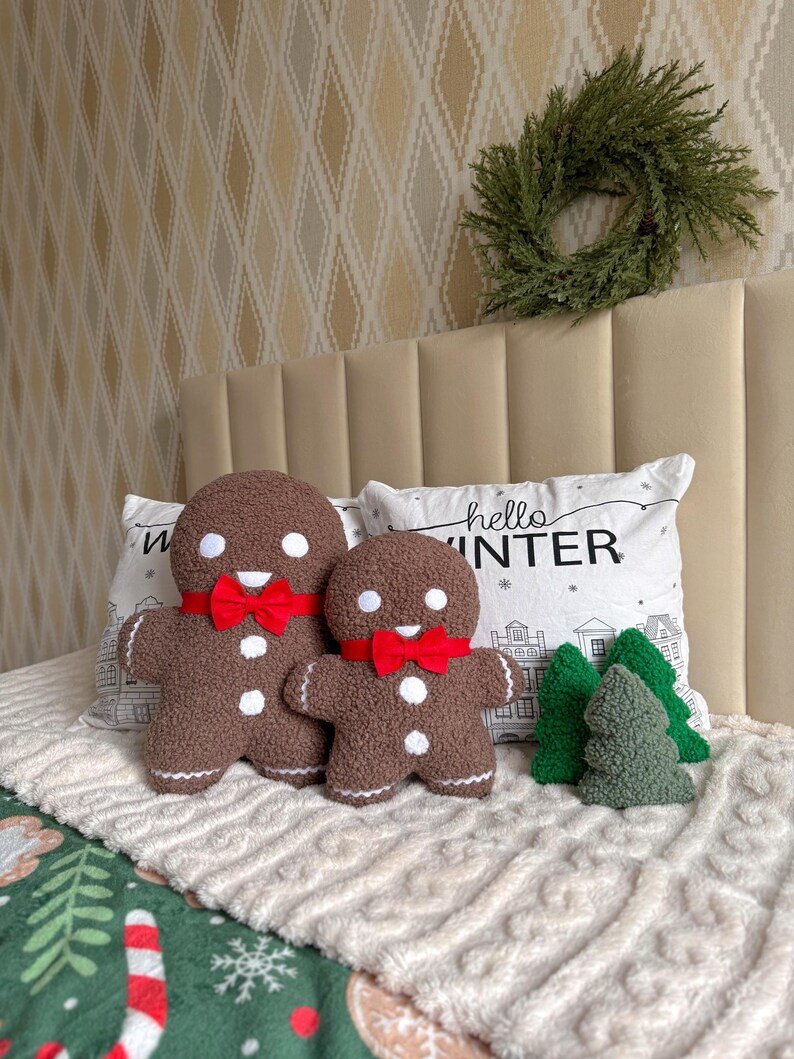 May include: Two brown textured gingerbread man plushies with white circular eyes and buttons, wearing red fabric bow ties. Beside them are three small green textured tree plushies. A white pillow with the text hello WINTER is behind them.