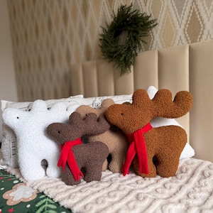 May include: A collection of four plush pillows shaped like reindeer and a bear. The set includes a white bear, a dark brown reindeer, a light brown reindeer, and a medium brown reindeer, each wearing a bright red scarf around its neck.