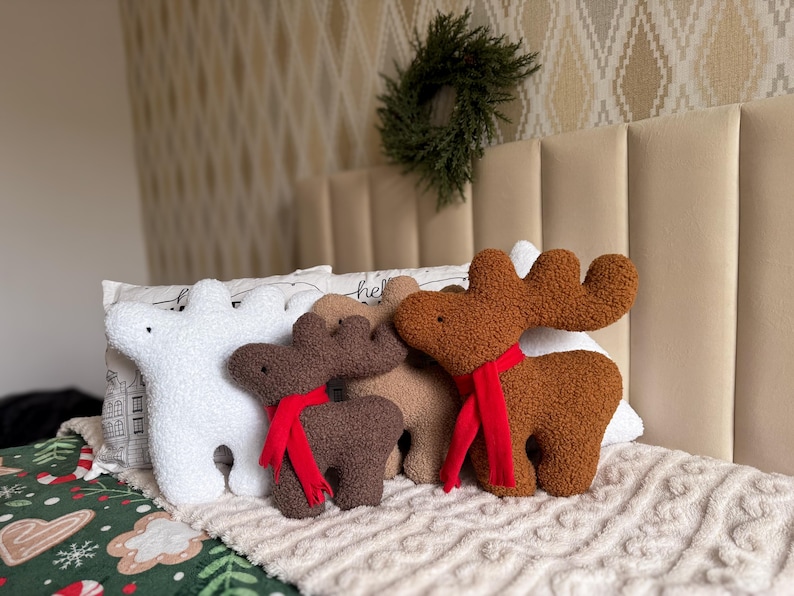 May include: A collection of four plush pillows shaped like reindeer and a bear. The set includes a white bear, a dark brown reindeer, a light brown reindeer, and a medium brown reindeer, each wearing a bright red scarf around its neck.
