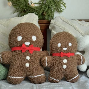 May include: Two gingerbread man plush toys with red bows, one large and one small, on a white couch with green and white pillows.