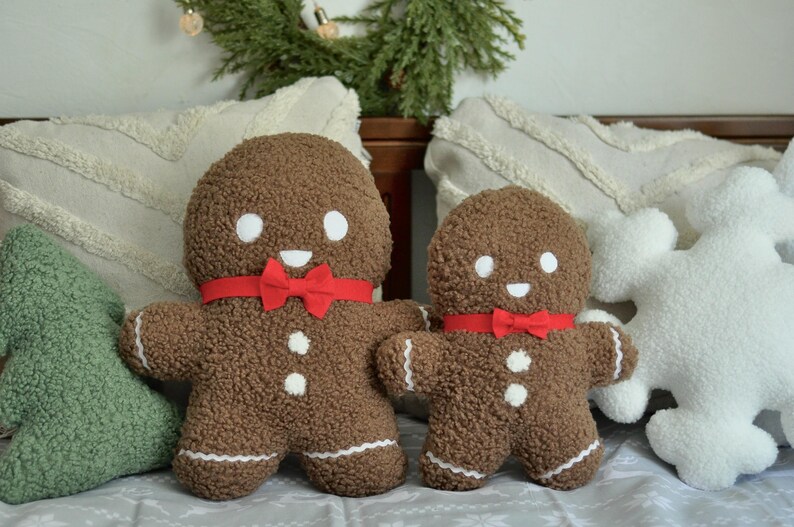 May include: Two gingerbread man plush toys with red bows, one large and one small, on a white couch with green and white pillows.