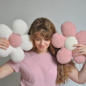 Può includere: Una donna tiene in mano due cuscini di peluche a forma di fiore. Un cuscino è bianco con un centro rosa, l'altro è rosa con un centro bianco.