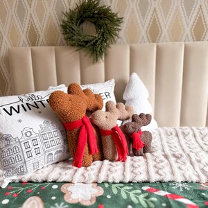 May include: Three plush reindeer figures in brown, tan, and dark brown, each wearing a bright red scarf. They are positioned in front of a white rectangular pillow with a black line drawing of buildings and the text "hello WINTER".