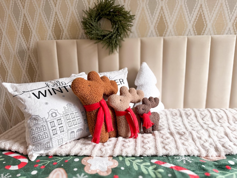 May include: Three plush reindeer figures in brown, tan, and dark brown, each wearing a bright red scarf. They are positioned in front of a white rectangular pillow with a black line drawing of buildings and the text "hello WINTER".