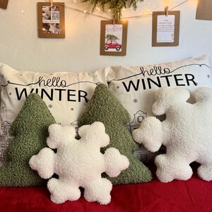 May include: Four decorative pillows with a winter theme. Two green pillows shaped like Christmas trees, one white pillow shaped like a snowflake, and one white pillow with black text that says "hello winter".