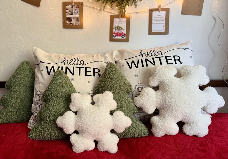 May include: Four decorative pillows with a winter theme. Two green pillows shaped like Christmas trees, one white pillow shaped like a snowflake, and one white pillow with black text that says "hello winter".