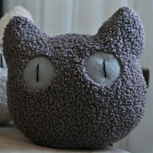May include: A dark gray cat-shaped pillow made of textured, curly fabric. The pillow features two pointed ears at the top and two large, round, light gray eyes with vertical black stitched pupils centered on the front of the plush object.