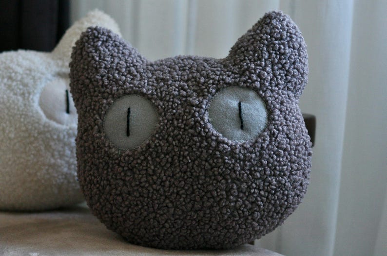 May include: A dark gray cat-shaped pillow made of textured, curly fabric. The pillow features two pointed ears at the top and two large, round, light gray eyes with vertical black stitched pupils centered on the front of the plush object.