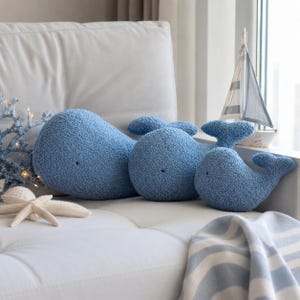 Peut inclure: Trois coussins en peluche en forme de baleine bleue avec une surface texturée et duveteuse et de petits yeux sombres. Les baleines sont disposées en rangée, diminuant de taille de gauche à droite, avec des corps arrondis et des nageoires caudales.