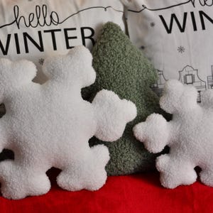 May include: Two white plush snowflake-shaped pillows, one larger than the other, are placed on a red surface. The pillows are surrounded by green and white pillows with the text "hello winter" printed on them.