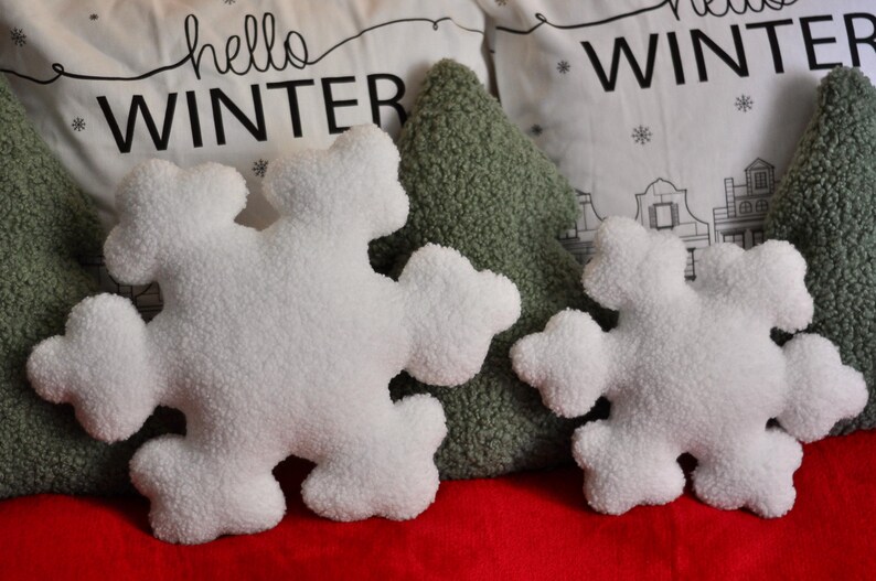 May include: Two white plush snowflake-shaped pillows, one larger than the other, are placed on a red surface. The pillows are surrounded by green and white pillows with the text "hello winter" printed on them.
