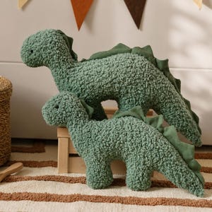May include: Two sage green dinosaur-shaped plush pillows made of textured sherpa fabric. Each dinosaur features a dark green fabric spine detail along the back, a rounded head, and a long tail. The plush toys are positioned one in front of the other.