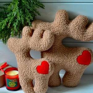 Puede incluir: Dos figuras de reno de peluche en color beige con textura tipo sherpa, cada una con un parche de coraz&oacute;n de fieltro rojo brillante en el costado. Los renos tienen astas distintivas y est&aacute;n dise&ntilde;ados como objetos decorativos suaves y rellenos.