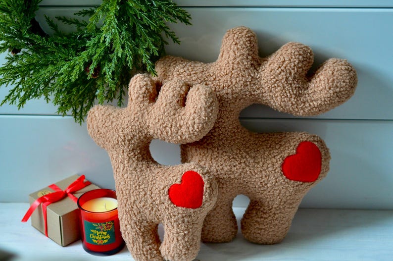 Puede incluir: Dos figuras de reno de peluche en color beige con textura tipo sherpa, cada una con un parche de coraz&oacute;n de fieltro rojo brillante en el costado. Los renos tienen astas distintivas y est&aacute;n dise&ntilde;ados como objetos decorativos suaves y rellenos.