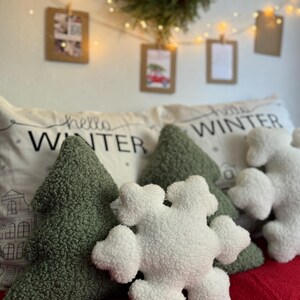 May include: Three plush pillows with winter-themed designs. A green tree-shaped pillow, a white snowflake-shaped pillow, and a white pillow with the text "hello winter" in black.