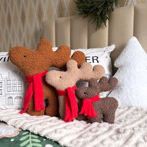 May include: Three plush reindeer-shaped pillows in varying shades of brown, each wearing a bright red scarf. The pillows have a textured, curly fabric finish. A white, tree-shaped pillow sits to the right, and a white pillow with black line art of buildings and the text "WINTER" is positioned behind the reindeer.