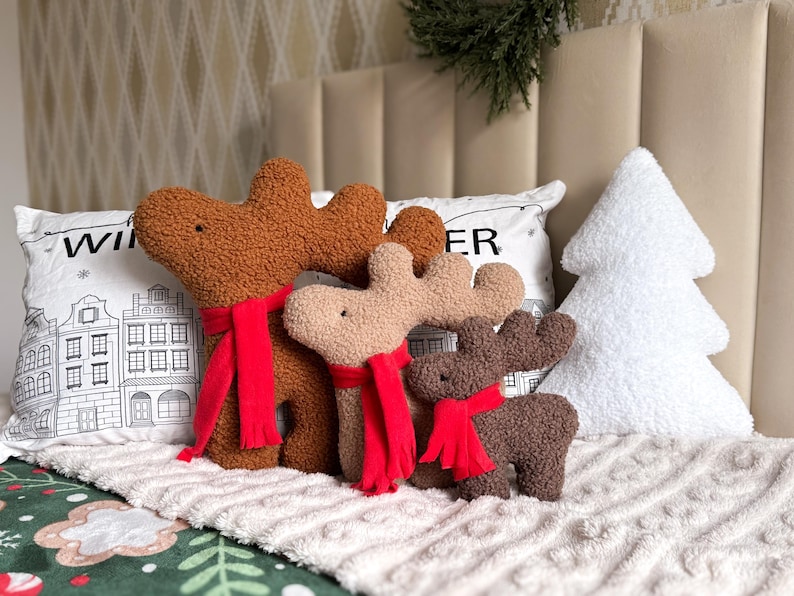 May include: Three plush reindeer-shaped pillows in varying shades of brown, each wearing a bright red scarf. The pillows have a textured, curly fabric finish. A white, tree-shaped pillow sits to the right, and a white pillow with black line art of buildings and the text "WINTER" is positioned behind the reindeer.