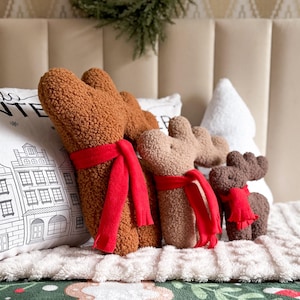 May include: Three plush reindeer figures made of brown textured sherpa fabric, each wearing a bright red scarf. The reindeer are positioned in front of a white pillow featuring a black line drawing of winter buildings and the text WINTER.