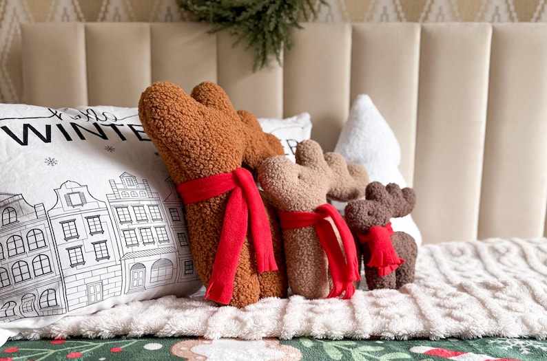 May include: Three plush reindeer figures made of brown textured sherpa fabric, each wearing a bright red scarf. The reindeer are positioned in front of a white pillow featuring a black line drawing of winter buildings and the text WINTER.