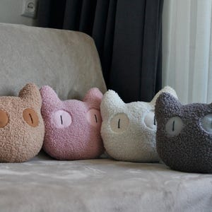May include: Four cat-shaped throw pillows made of textured, fuzzy fabric. The pillows are arranged in a row and come in four distinct colors: tan, light pink, off-white, and dark gray. Each pillow features two large, oval-shaped eyes.