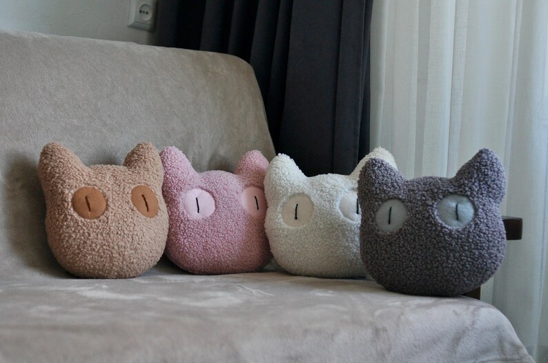 May include: Four cat-shaped throw pillows made of textured, fuzzy fabric. The pillows are arranged in a row and come in four distinct colors: tan, light pink, off-white, and dark gray. Each pillow features two large, oval-shaped eyes.