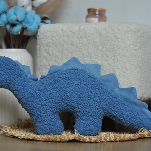 May include: A blue dinosaur-shaped plush toy made of textured sherpa fabric. The dinosaur features a long neck, a rounded head with a small dark eye, and a row of triangular spikes running along its back down to the tail. The plush has a soft, fuzzy appearance and a solid blue color throughout its body.