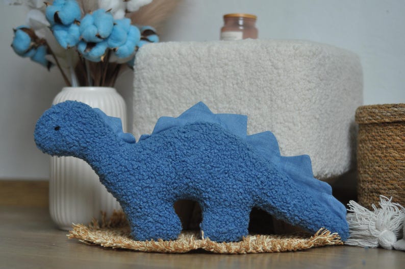 May include: A blue dinosaur-shaped plush toy made of textured sherpa fabric. The dinosaur features a long neck, a rounded head with a small dark eye, and a row of triangular spikes running along its back down to the tail. The plush has a soft, fuzzy appearance and a solid blue color throughout its body.