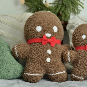 May include: Two gingerbread man plush toys with red bow ties. The larger gingerbread man is brown with white icing details and a smiling face. The smaller gingerbread man is brown with white icing details and a smiling face. The gingerbread men are sitting on a white surface.