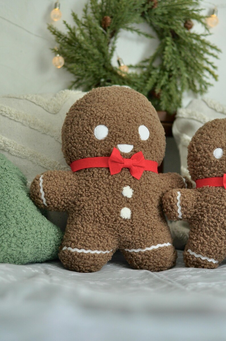 May include: Two gingerbread man plush toys with red bow ties. The larger gingerbread man is brown with white icing details and a smiling face. The smaller gingerbread man is brown with white icing details and a smiling face. The gingerbread men are sitting on a white surface.