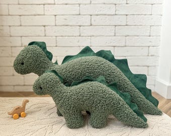 May include: Two plush dinosaur toys made of olive green textured sherpa fabric with dark green felt spines along their backs. A small wooden dinosaur toy with yellow wheels is positioned to the left of the two larger plush dinosaurs.